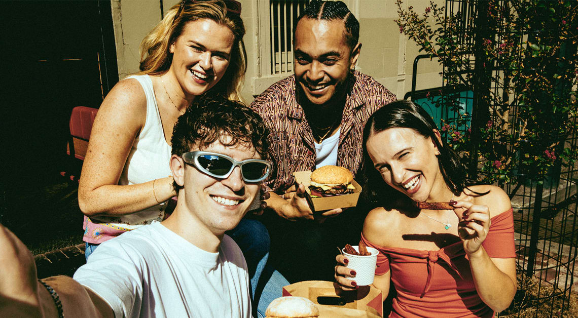A group of friends sits outside at a restaurant, enjoying food, with a man and woman holding cheeseburgers and fries.